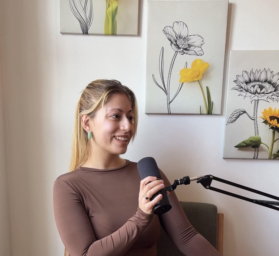 Annie sitting in a green armchair, smiling and holding a professional podcast microphone in front of a wall with floral artwork.